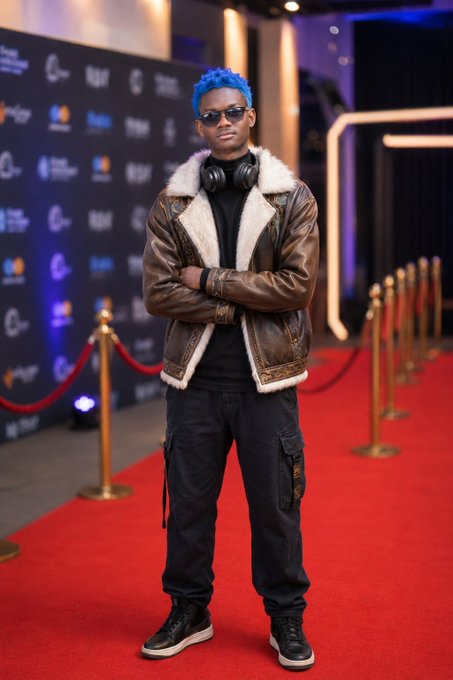 A man with bright blue hair and sunglasses stands on a red carpet wearing a brown shearling jacket, black turtleneck, and headphones around his neck.