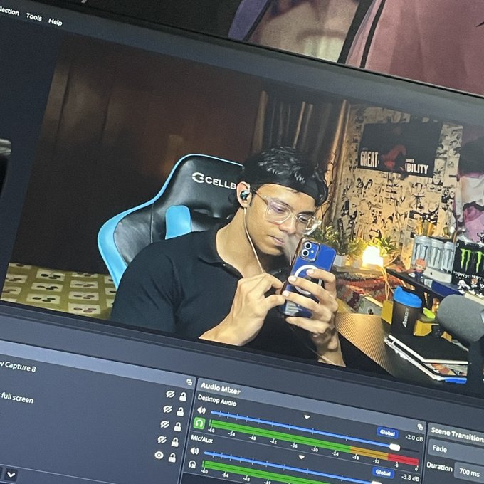 A computer monitor displaying live streaming software (OBS) with an Audio Mixer panel, showing a young man wearing glasses and earbuds sitting in a gaming chair using his smartphone, with a Monster Energy poster in the background.