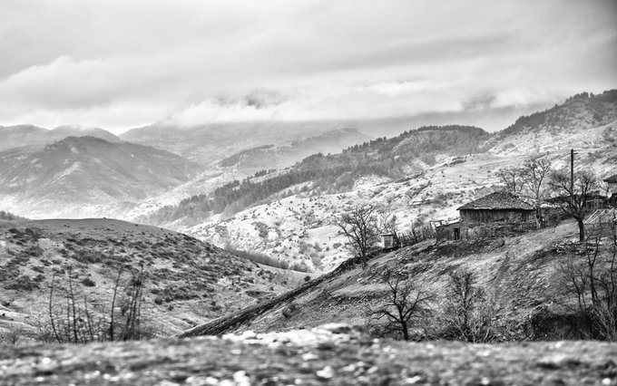 A black and white landscape photograph depicting a remote stone house on a hillside in a mountainous, snow-patched terrain under cloudy skies.