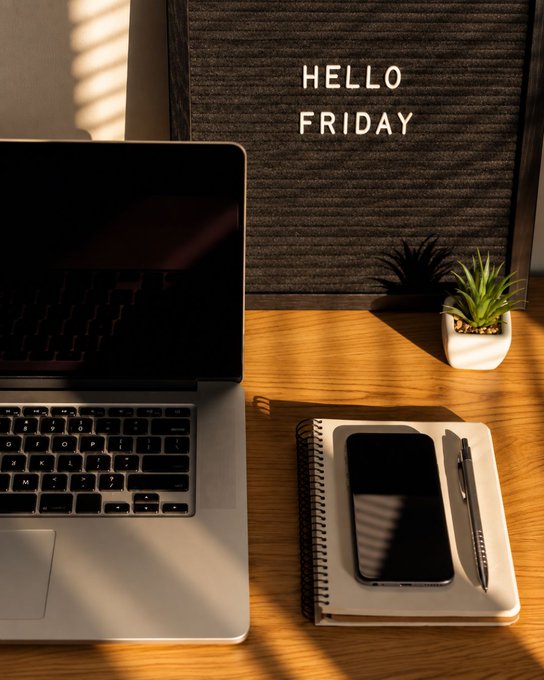A workspace scene with a laptop, smartphone, and pen on a notebook next to a small plant, with a letter board displaying 'HELLO FRIDAY' in warm sunlight.