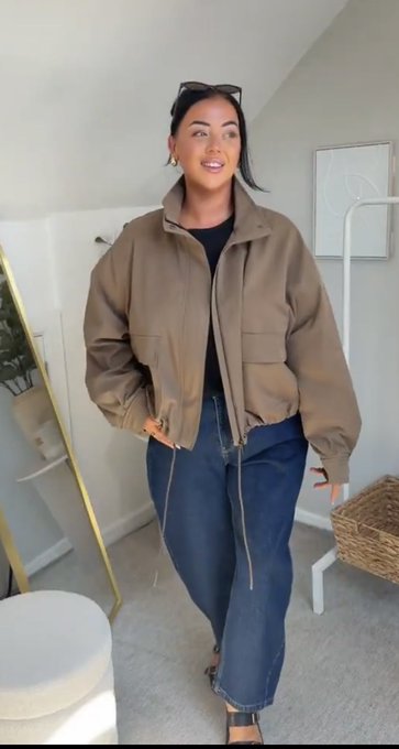 A woman models a brown zip-up jacket paired with baggy blue jeans, sunglasses resting on her head, standing in a bright room.