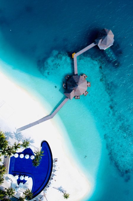 Aerial photograph of a tropical resort featuring turquoise ocean water, a white sand beach, a wooden walkway connecting overwater bungalows, palm trees, and a heart-shaped blue swimming pool.