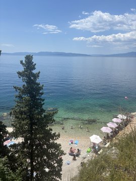 An elevated view of a beach scene with clear turquoise water, a sandy shore dotted with white umbrellas, a tall pine tree in the foreground, and mountains visible in the distance under a partly cloudy blue sky.