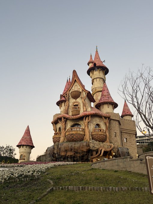 A whimsical fairytale-style castle with turrets, red roofs, balconies, and large root-like supports stands above landscaped flowerbeds.