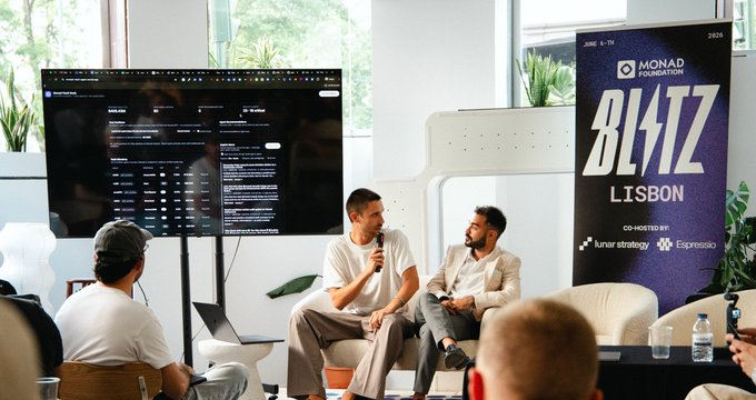 The image shows a group of people sitting around a table with laptops open in front of them. There are two men seated at the table, one wearing a white shirt and the other wearing a black jacket. They appear to be engaged in conversation or working on their projects. The room has a large screen mounted on the wall behind them, which could be used for presentations or discussions.

In addition to the people sitting around the table, there are several chairs placed throughout the room. A potted plant is visible near one of the chairs, and multiple bottles can be seen scattered across the floor. The overall atmosphere suggests a collaborative environment where individuals come together to work on projects or share ideas.