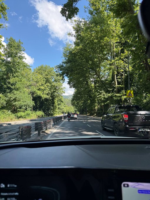 A view from inside a car driving down a tree-lined road with a motorcyclist visible ahead on a sunny day.