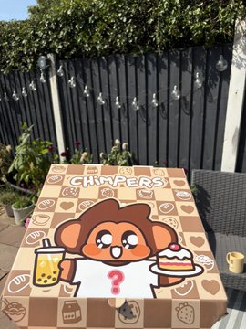 An outdoor patio table covered with a themed cloth featuring a cartoon monkey character surrounded by pastries, with the text 'CHIMPERS' against a dark fence background.