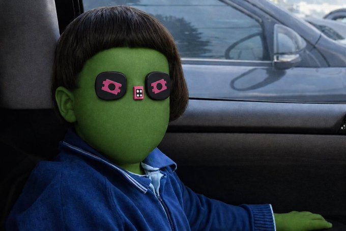 A stylized, AI-edited portrait of a child with a green alien-like face and pink robotic eyes, wearing a blue zip-up jacket and sitting in a car with another vehicle visible through the window.