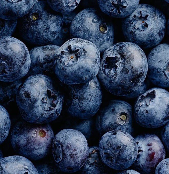A close-up macro photograph shows a pile of fresh blueberries, highlighting their dusty blue skin and star-shaped calyx crowns.