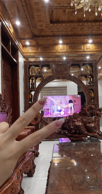 A hand makes a peace sign in front of an ornate wooden living room featuring a large TV displaying a pink-hued scene, where a person stands in a bedroom with another figure in white. The caption connects to the post's hopeful sentiment, suggesting the peace sign symbolizes optimism and anticipation for good news upon waking.