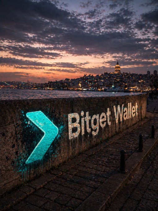 A "Bitget Wallet" logo projected onto a stone wall by the water in Istanbul at dusk, with the Galata Tower and illuminated cityscape in the background. The image reinforces the post's message by visually placing Bitget Wallet at the crossroads of a storied city, symbolizing its role as a gateway to new Web3 narratives.