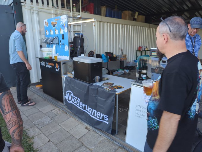 An outdoor beer festival scene featuring a "Moderunners" table with beer taps, where a man in a black shirt holds a glass of beer while others stand around the serving area.