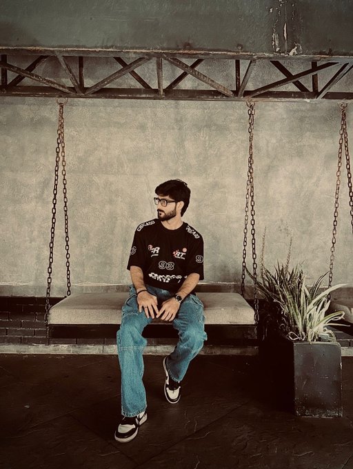 A man with glasses and a beard, wearing a black graphic t-shirt and blue jeans, sits on a suspended swing bench in an industrial-style interior with concrete walls and chains.