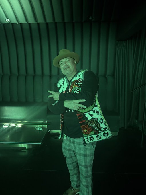 A person wearing a tan cowboy hat, a patterned vest over a black long-sleeve shirt, and plaid pants, posing with crossed arms in a room illuminated by green light.
