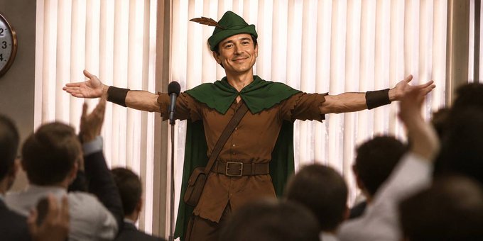 A man dressed in a Robin Hood costume—complete with a green feathered cap, cape, and brown tunic—stands triumphantly before a microphone with his arms outstretched, smiling broadly as an applauding crowd surrounds him.