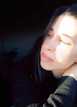 A selfie of a young woman with dark hair resting her head on her hand, with warm sunlight illuminating half of her face.
