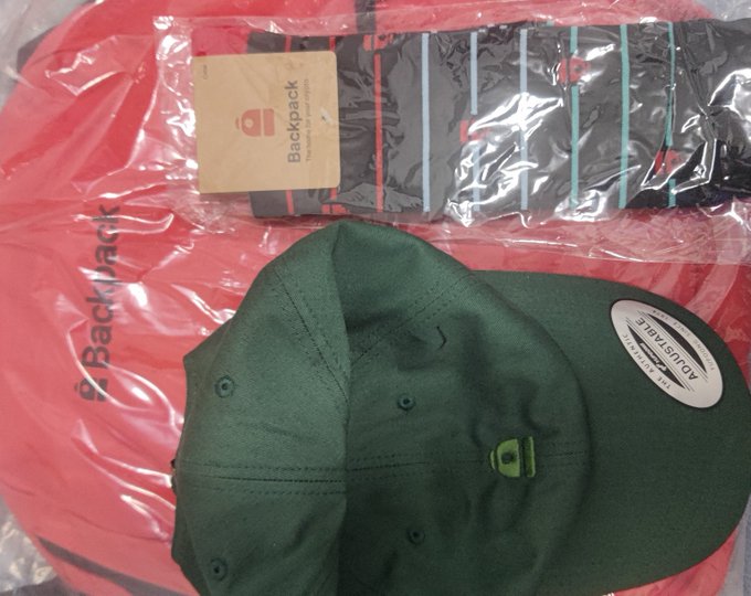 A green 'Backpack' branded cap with an 'ADJUSTABLE' tag resting on a red plastic bag alongside another packaged item.