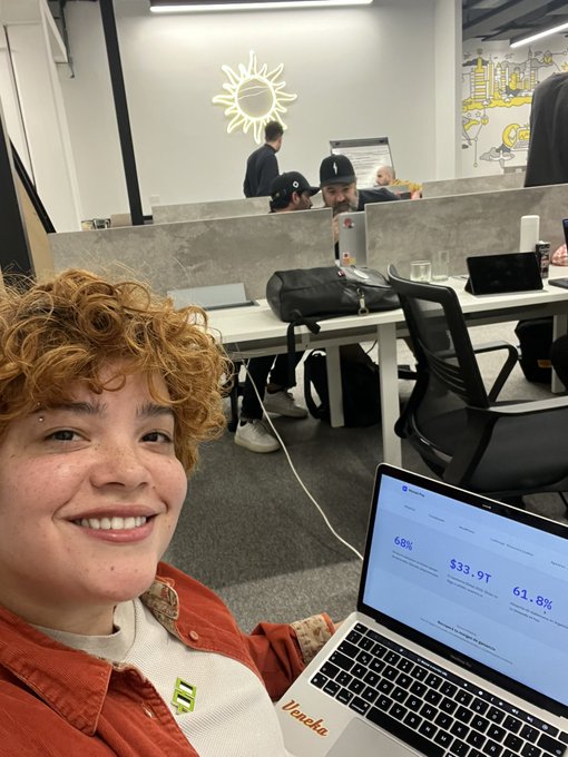 In this image, there are multiple people present in a brightly lit room with white walls and a gray floor. A young girl wearing an orange shirt is sitting down at a desk, smiling as she holds up her laptop computer displaying various statistics about ecommerce sales. She has tagged someone on Instagram to accompany her in the photo. The rest of the image features more people scattered throughout the space, engaging in their own activities or observing the scene. A sunburst light and some text are also visible in the background.