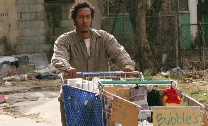 A man with messy hair pushes a shopping cart filled with cardboard boxes labeled "Bubbles" through a cluttered street, capturing the disheveled, desperate look of a meme coin shiller who DM's random tickers to strangers. The image humorously embodies the chaotic energy of someone relentlessly promoting low-value crypto projects to anyone who will listen.