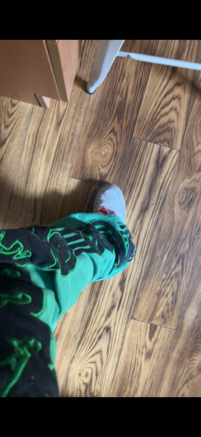 A first-person view looking down at a leg wearing green patterned pants and a white sock, standing on a wooden floor.
