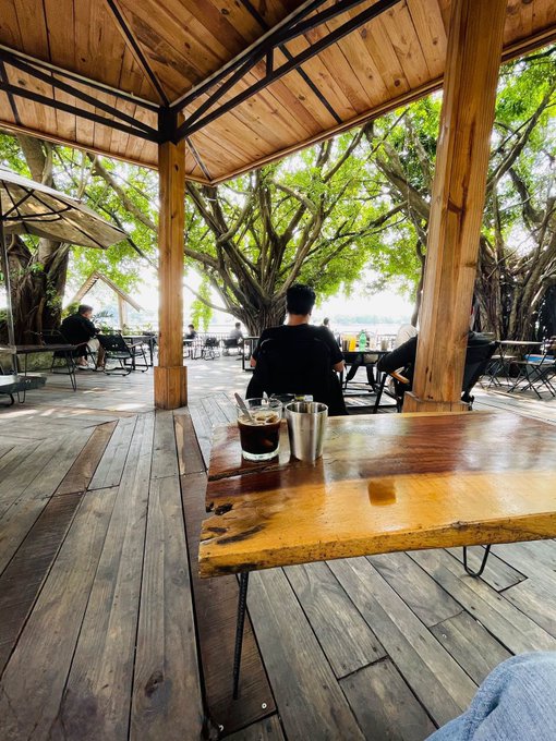 An outdoor café or restaurant with a wooden roof overlooking trees and water, featuring a wooden table in the foreground with a glass of dark beverage, a metal tumbler, and salt and pepper shakers.
