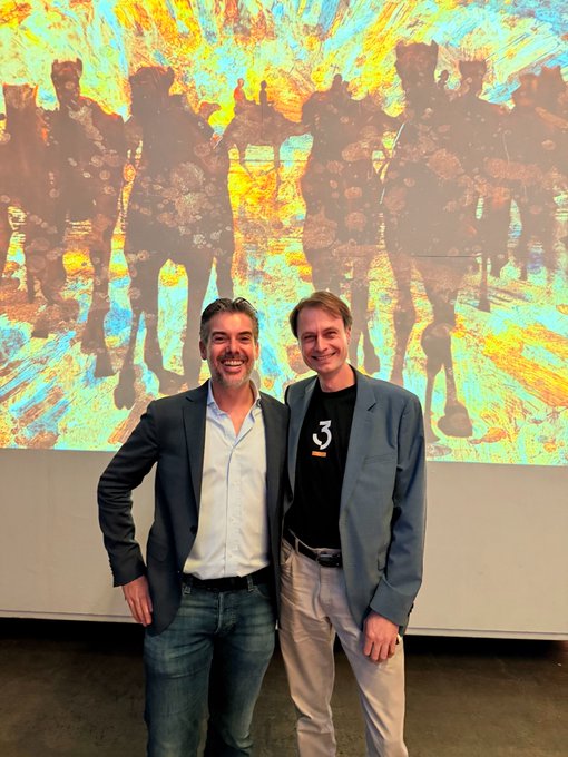 Two men smile and stand in front of a colorful abstract painting depicting silhouetted figures on horseback.