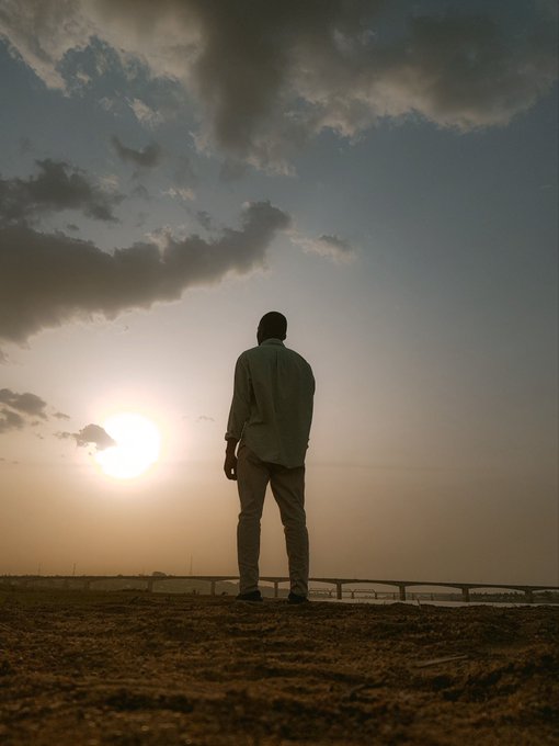 A silhouetted man stands on a sandy beach facing a sunset over the water under a cloudy sky.