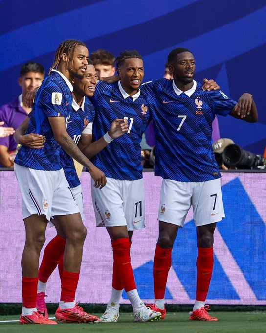 Four French national football team players in blue jerseys and white shorts, including number 7 and 11, celebrating together on the pitch with arms around each other.