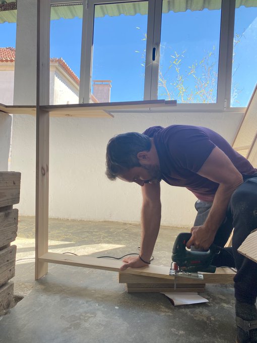 A man in a dark t-shirt uses a jigsaw to cut a piece of wood on the floor of what appears to be a patio or covered porch area, with large windows behind him.
