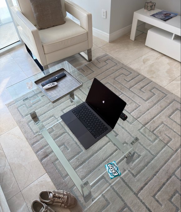 A laptop sits open on a glass coffee table in a living room, with sneakers placed on the floor nearby and a pack of Extra chewing gum resting on the table's edge.