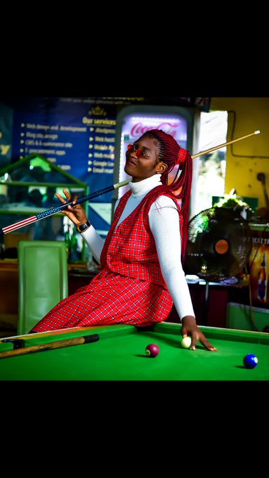 The image features a woman sitting at a pool table, holding two pool cues while wearing a red dress. The background includes a green chair and a yellow sign with white text that reads "coke".