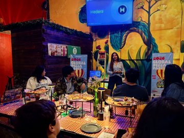 The image shows a group of people gathered around tables, enjoying pizza together. There are at least six visible individuals seated at various dining tables with plates of food in front of them. The room has a colorful mural on one wall that adds vibrancy to the atmosphere. A woman is standing behind a table and giving a presentation or speech, possibly about global warming or another topic related to the event. The image also includes text elements such as "En estos días he estado aprendiendo más sobre algunas novedades de @Hedera como CLPR, HashSphere y el tema de AI agents y justo para el Pizza Day en Puebla me puse a estudiar un poco más y pude platicar de esto con la comunidad."
