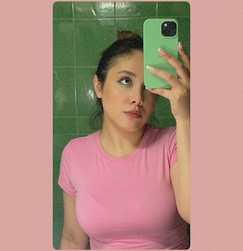 A woman in a pink t-shirt takes a mirror selfie with a green phone case against a green tiled wall, matching the soft pink and green palette of her "The Quiet Between" NFT auction drop starting at 0.111 ETH. The minimalist bathroom aesthetic complements the understated, introspective vibe of her Thursday artist feature.