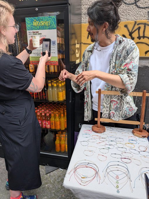 The image shows a woman taking a picture of another woman holding up a bracelet, which appears to be made from beads or strings. The scene takes place outside a vending machine that sells drinks, with a sign above it advertising "Day 4: Wow Berlin you've been amazing. We've seen the Blockspäti vendors learn and then accept their first $ZEC sales all in one day." This event is about promoting the sale of these beverages to raise awareness for a charity or cause related to theBlockspäti vendors, who are learning how to sell their products effectively.
