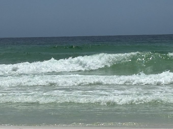 Ocean waves crash onto a sandy shore under a clear blue sky, with a few small figures visible surfing or swimming in the surf. The image evokes the outdoor, nature-centered lifestyle the post promotes—suggesting that living near the sea and embracing natural elements like sun and sea air (referencing the Lenard effect, which describes the negative electrical charge produced by water breaking) offers a path to health independent of conventional medicine.