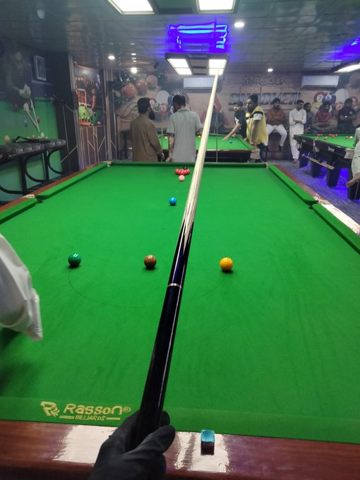 First-person view from behind a cue stick on a green Rasson billiards table with several colored balls, looking toward other players in a pool hall with blue neon lighting.
