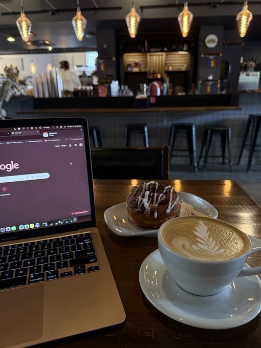 A laptop displaying a Google search page sits on a wooden café table beside a latte with leaf latte art and a glazed pastry, with pendant lights and bar stools visible in the background.