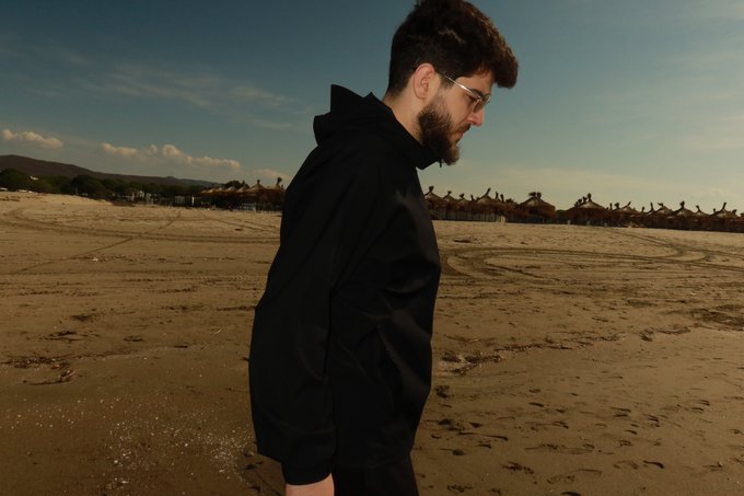 A bearded man wearing glasses and a black jacket stands in profile on a sandy beach with thatched-roof structures in the background.