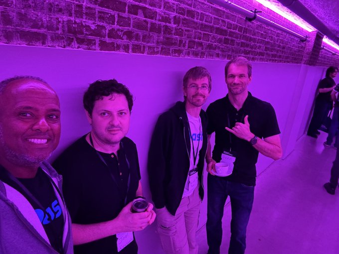 A purple-tinted selfie of four men posing together at what appears to be a crypto or tech conference, with two making hand gestures and all wearing lanyards.