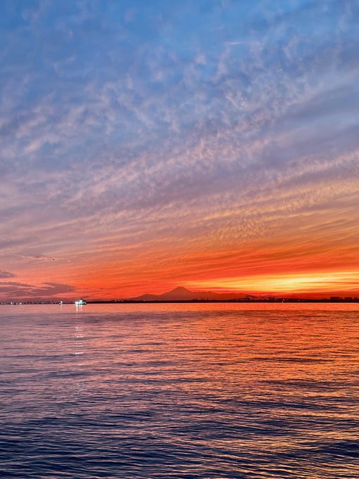 A serene sunset over a calm body of water with vibrant orange and red hues blending into a blue cloudy sky, with distant mountains on the horizon.