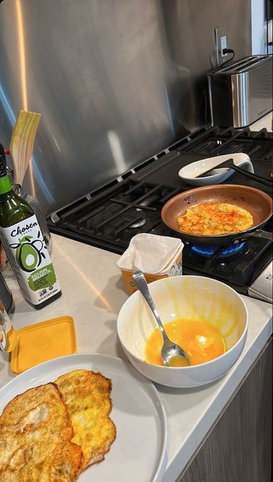 A kitchen scene shows a frying pan with cooking eggs on a gas stove, alongside a bowl of beaten eggs, a Chosen avocado oil bottle, and a plate of finished omelets.