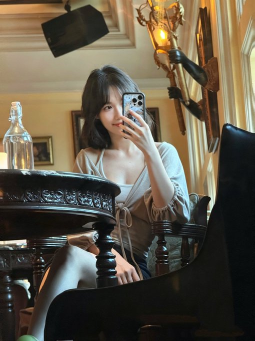A young woman with dark hair taking a mirror selfie with her phone while seated at an ornate dark wooden table, with a glass bottle nearby and decorative wall lamps in the background.