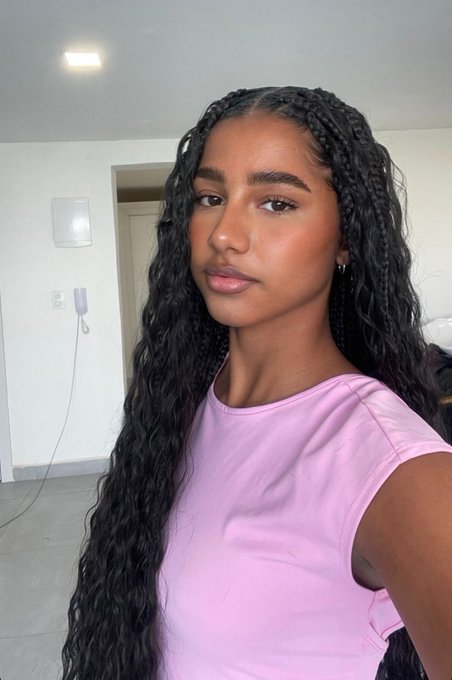 A selfie of a young woman with long dark braided hair wearing a pink sleeveless top, posing indoors in front of a white wall.