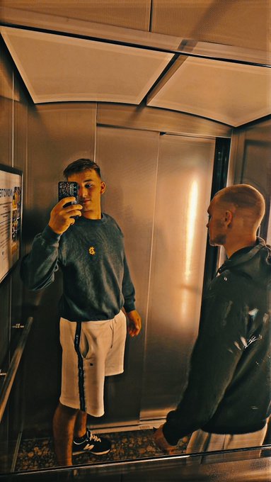 A mirror selfie taken inside an elevator showing a young man in a blue sweatshirt and white shorts holding a phone, with another person standing beside him against the metallic elevator walls.