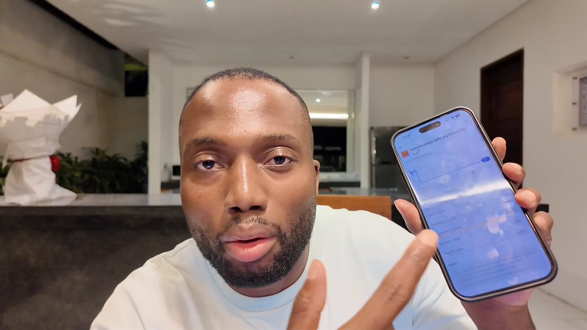 The image shows a man holding up his phone, which displays an app called "Strategy". The man appears to be posing for a photo or video, possibly sharing information about the app with others. The background of the image features a dining table surrounded by chairs and potted plants, suggesting that this scene takes place in someone's home or office setting.