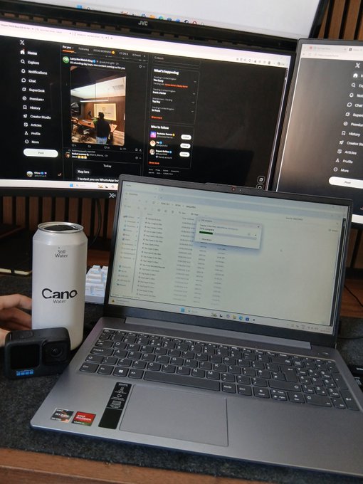 A desk setup featuring a laptop displaying a file transfer dialog box, positioned in front of two monitors showing social media feeds, with a white "Cano Water" can and a GoPro camera nearby.