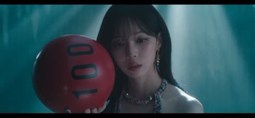 A dark-haired woman poses with a red ball marked '100' while wearing a sparkling necklace in a dimly lit, bluish setting.