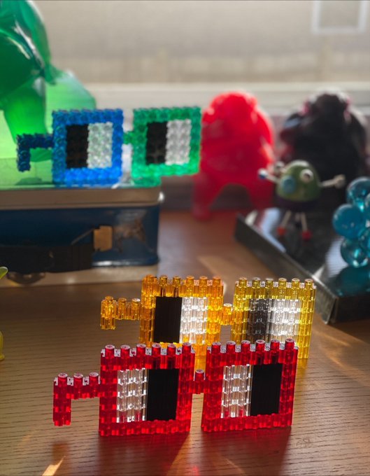 Colorful pixel art creations made from Perler beads sit on a wooden surface, featuring blocky character designs in red, yellow, blue, and green, with toy figures in the background.