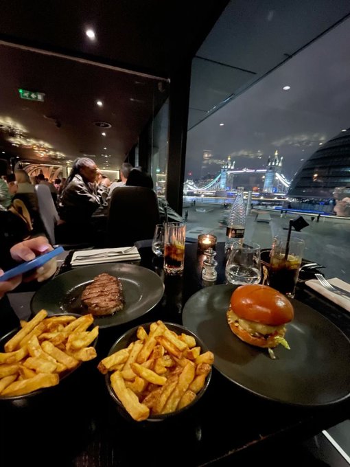 A restaurant dinner featuring a steak, burger, and two baskets of fries sits on a table by the window, with Tower Bridge illuminated in the background at night, ready to fuel a long charting session.