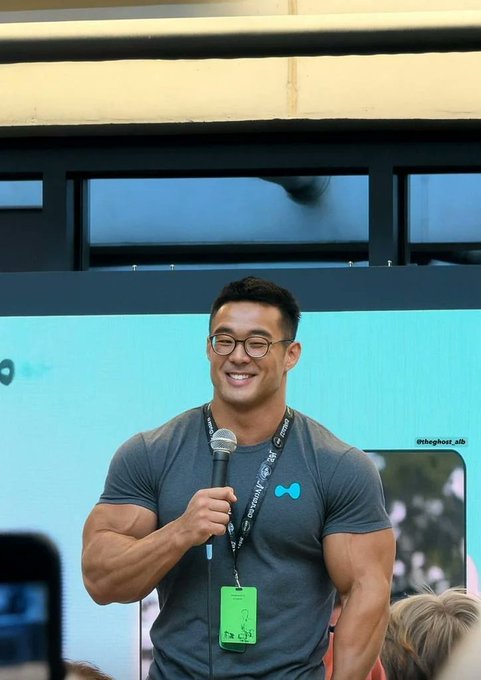 A smiling muscular man wearing glasses and a gray t-shirt, holding a microphone while standing in front of a screen displaying '@itsghost_adb'.